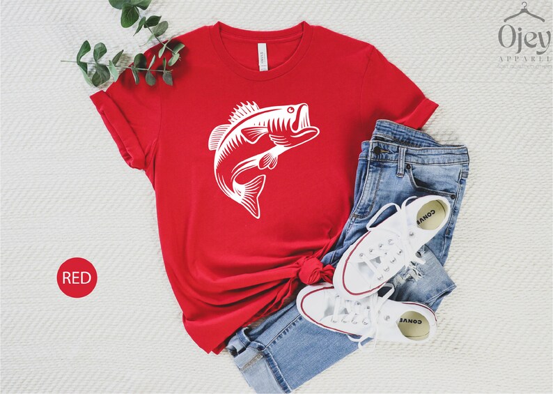 May include: Red t-shirt featuring a white graphic of a bass fish leaping out of the water. The shirt is laid out on a white surface with a pair of white trainers and blue jeans.