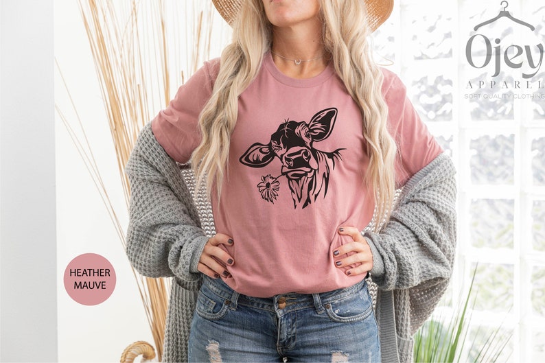 May include: A woman wearing a heather mauve t-shirt with a black silhouette of a cow with a flower in its mouth. The t-shirt is a relaxed fit and has a crew neck. The woman is also wearing a gray cardigan and blue jeans.