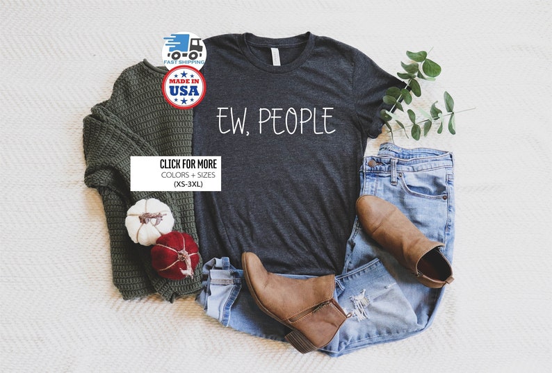 May include: A dark heather grey t-shirt with the text "EW, PEOPLE" in white lettering. The shirt is laid flat on a white surface with a green sweater, jeans, and brown boots.