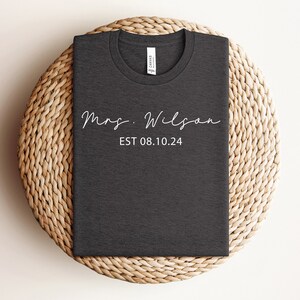 May include: A dark gray t-shirt with the text "Mrs. Wilson EST 08.10.24" in white.