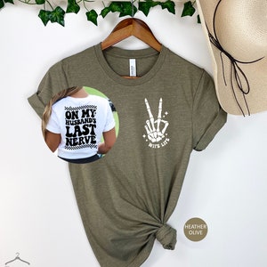 May include: A green t-shirt with a white skeleton hand making a peace sign and the text "Wife Life". The back of the shirt has the text "On My Husband's Last Nerve" in black and white.