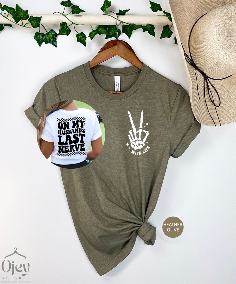 May include: A green t-shirt with a white skeleton hand making a peace sign and the text "Wife Life". The back of the shirt has the text "On My Husband's Last Nerve" in black and white.