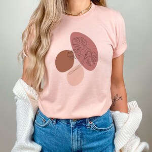 Puede incluir: Una camiseta rosa con un dise&ntilde;o gr&aacute;fico de formas abstractas y una hoja en colores marr&oacute;n, rosa y rojo.