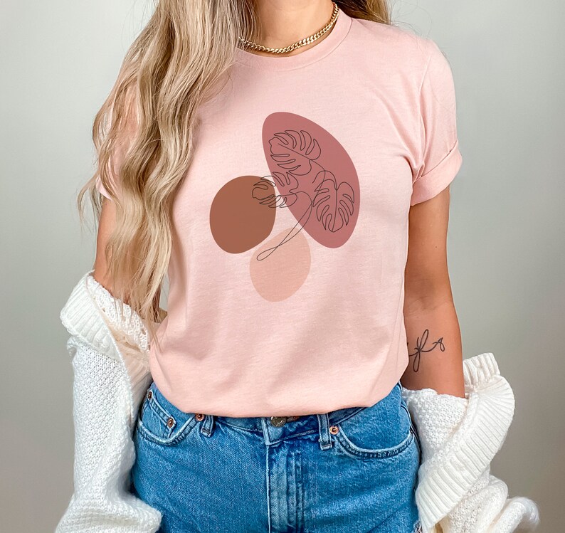 Puede incluir: Una camiseta rosa con un dise&ntilde;o gr&aacute;fico de formas abstractas y una hoja en colores marr&oacute;n, rosa y rojo.