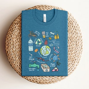 May include: A teal blue t-shirt with a graphic design of various ways to help save the planet. The graphic includes a bicycle, a lightbulb, a water bottle, a recycling symbol, a turtle, a tree, and the words "save the planet", "don't litter", "save water", "save life", "plant a tree", "recycle", "reduce", "reuse", "skip a straw", "turn off lights", "bike more drive less", "save a turtle", "upcycle things", and "volunteer".