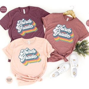 May include: Three t-shirts with the text "Third Grade" in a retro rainbow font. The shirts are in heather maroon, heather peach, and heather mauve.