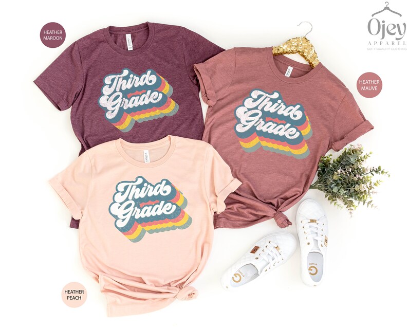 May include: Three t-shirts with the text "Third Grade" in a retro rainbow font. The shirts are in heather maroon, heather peach, and heather mauve.