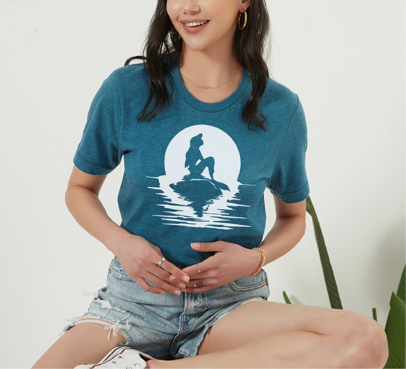 K&ouml;nnte beinhalten: Ein blaues T-Shirt mit einem wei&szlig;en Silhouettenbild von Arielle, der kleinen Meerjungfrau, die auf einem Felsen vor einem Vollmond sitzt. Der Mond spiegelt sich im Wasser darunter.