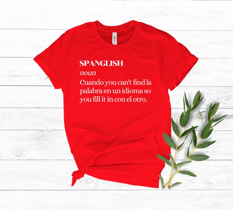 Puede incluir: Camiseta roja con texto blanco que dice "SPANGLISH noun Cuando you can't find la palabra en un idioma so you fill it in con el otro."
