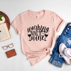 May include: A pink t-shirt with the text "working nine to wine" in black lettering. The shirt is styled with a pair of blue jeans and white sneakers.
