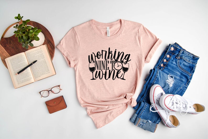 May include: A pink t-shirt with the text "working nine to wine" in black lettering. The shirt is styled with a pair of blue jeans and white sneakers.