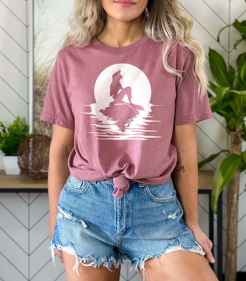 K&ouml;nnte beinhalten: Ein mauvefarbenes T-Shirt mit einer wei&szlig;en Silhouette einer Meerjungfrau, die auf einem Felsen vor einem Vollmond sitzt. Der Mond spiegelt sich im Wasser darunter.