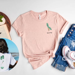 May include: A pink t-shirt with a green hand graphic and the text "Rise to Glory". The shirt is shown with a pair of blue jeans and white sneakers.