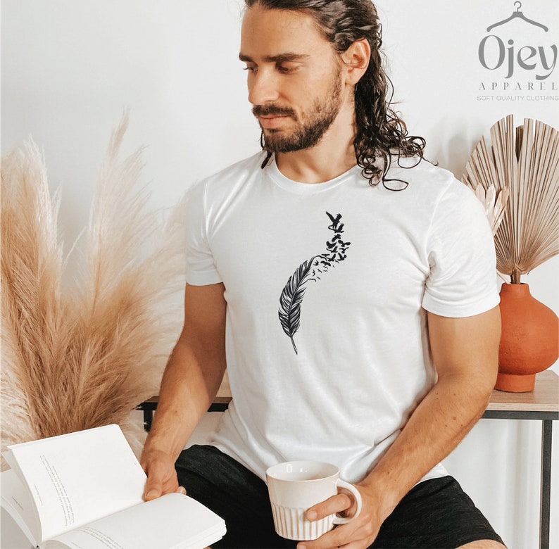 Op de afbeelding: Een wit T-shirt met een zwart-witte afbeelding van een veer met vogels die er vanaf vliegen. De afbeelding is gecentreerd op het T-shirt.