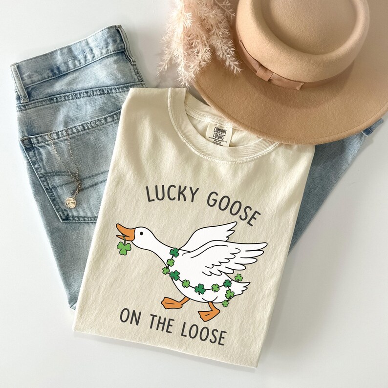 May include: Cream-colored t-shirt with the text "LUCKY GOOSE ON THE LOOSE" and a cartoon goose wearing a shamrock garland. The shirt is paired with light-wash jeans, a tan hat, and a necklace.
