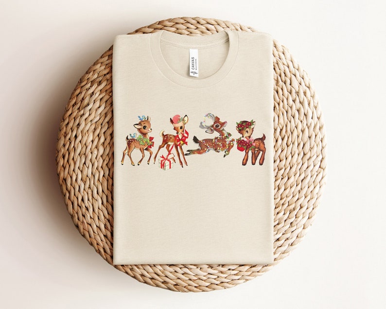 Pu&ograve; includere: Una maglietta beige con un'illustrazione vintage di quattro cervi che indossano cappelli di Natale e tengono dei regali. I cervi sono marroni con macchie bianche e hanno fiocchi rossi intorno al collo.