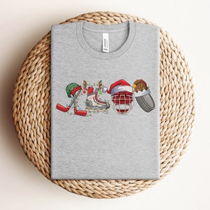 May include: Gray t-shirt with a Christmas-themed hockey design. The design features a hockey stick, a hockey skate, a hockey helmet, and a hockey puck, all decorated with Christmas lights and ornaments.