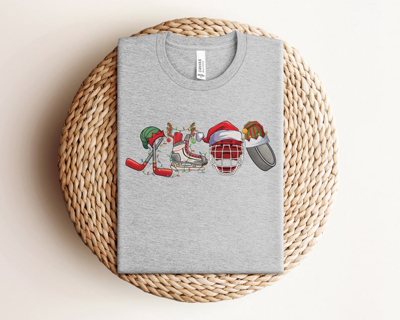 May include: Gray t-shirt with a Christmas-themed hockey design. The design features a hockey stick, a hockey skate, a hockey helmet, and a hockey puck, all decorated with Christmas lights and ornaments.