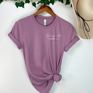 May include: Mauve t-shirt with rolled sleeves, hanging on a wooden hanger. The shirt displays the text "Smiles for all - DENTAL" in white script. A straw hat is visible in the background.