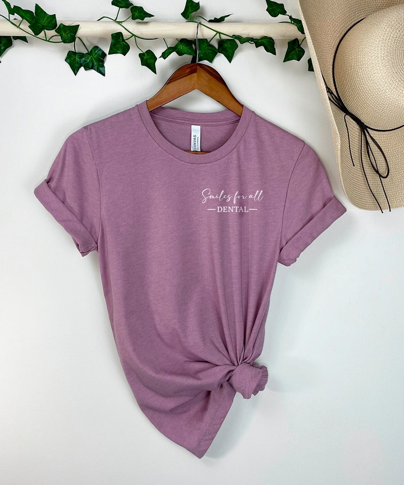 May include: Mauve t-shirt with rolled sleeves, hanging on a wooden hanger. The shirt displays the text "Smiles for all - DENTAL" in white script. A straw hat is visible in the background.