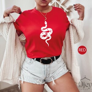 May include: Red t-shirt with a white graphic of a snake with stars and moons. The snake is curled up and has its head facing the right. The t-shirt is being worn by a person. The word "RED" is written in white letters on a red background in the bottom right corner of the image.