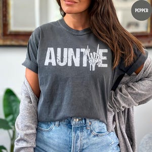 May include: Dark grey t-shirt with the word "AUNTIE" in distressed white lettering. The "E" is replaced with a white skeleton hand making a peace sign. The shirt is worn with blue jeans and a grey cardigan.