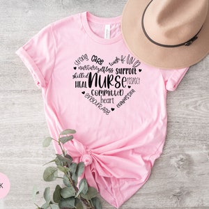 May include: Pink t-shirt with a black graphic design that says "Strong, Care, Tough, Kind, Nurture, Selfless, Support, Skilled, Nurse, Respect, Heal, Committed, Heart, Encourage, Compassion".