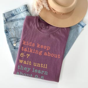 May include: A mauve t-shirt with the text "kids keep talking about 6-7 wait until they learn about 9-5" in orange, yellow, and teal. The shirt is paired with light blue jeans and a beige hat.