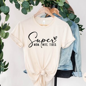 May include: A beige t-shirt with the text "Super Mom. Wife. Tired." in black cursive lettering and a small heart.