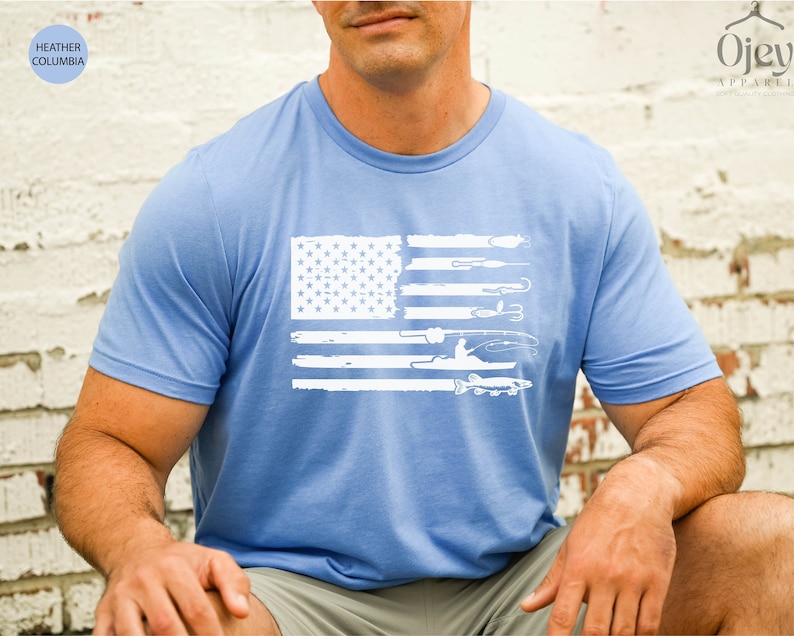 K&ouml;nnte beinhalten: Ein hellblaues T-Shirt mit einem wei&szlig;en, abgenutzten Design der amerikanischen Flagge. Die Flagge zeigt fischereibezogene Bilder, darunter einen Fischer in einem Boot, Angelruten, Haken und Fische.