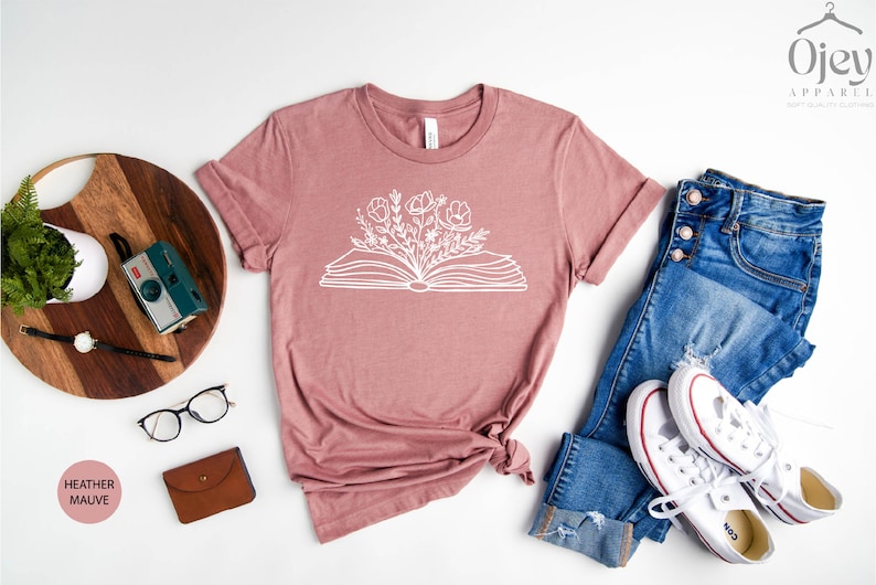 K&ouml;nnte beinhalten: Ein T-Shirt in Heide-Mauve mit einer wei&szlig;en Grafik eines offenen Buches, aus dem Blumen wachsen. Das Shirt liegt flach auf einer wei&szlig;en Oberfl&auml;che mit einer Jeans, einem Paar wei&szlig;en Sneakers, einem Holzbrett, einer Topfpflanze, einer Kamera und einer Brille.