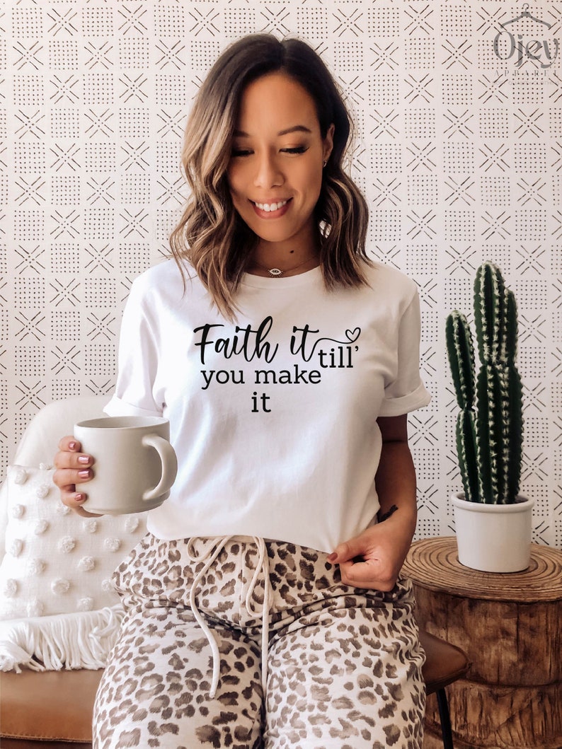 K&ouml;nnte beinhalten: Wei&szlig;es T-Shirt mit dem Text "Faith it till' you make it" und einem kleinen Herzsymbol. Die Person, die das Shirt tr&auml;gt, sitzt auf einem Sofa und h&auml;lt eine wei&szlig;e Tasse.