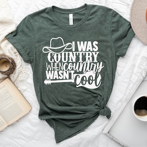 May include: A dark green t-shirt with a white graphic that says "I was country when country wasn't cool". The graphic also includes a cowboy hat and a guitar.