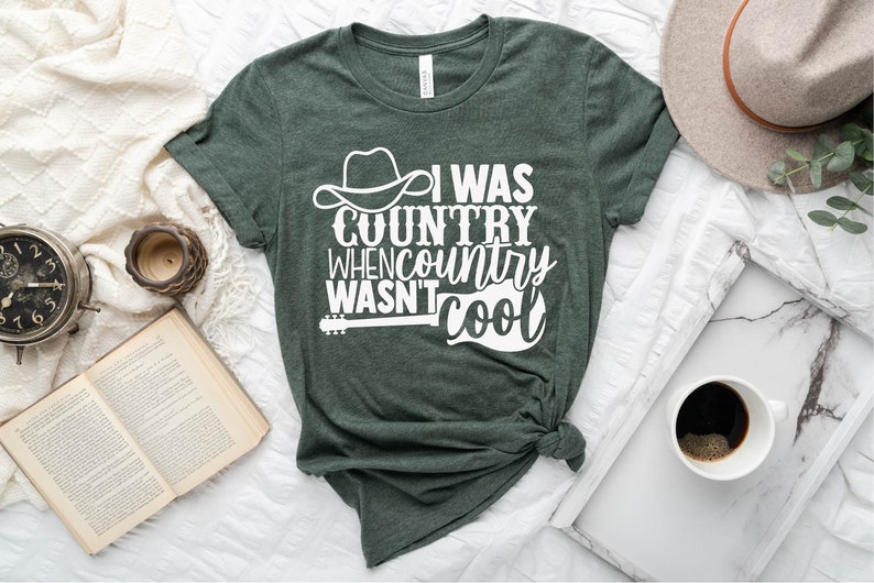 May include: A dark green t-shirt with a white graphic that says "I was country when country wasn't cool". The graphic also includes a cowboy hat and a guitar.