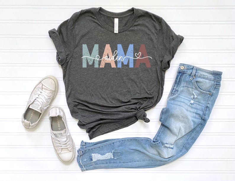 May include: Dark gray t-shirt with the word "MAMA" in colorful block letters and the name "Aiden" in script. The shirt is paired with light blue jeans and white sneakers, creating a casual, everyday outfit. A small heart is next to the name.