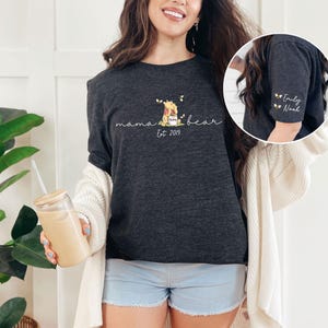 May include: A dark grey t-shirt with the text "mama bear est. 2019" and a small image of Winnie the Pooh holding a jar of honey. The sleeves have the names "Emily" and "Noah" embroidered on them.