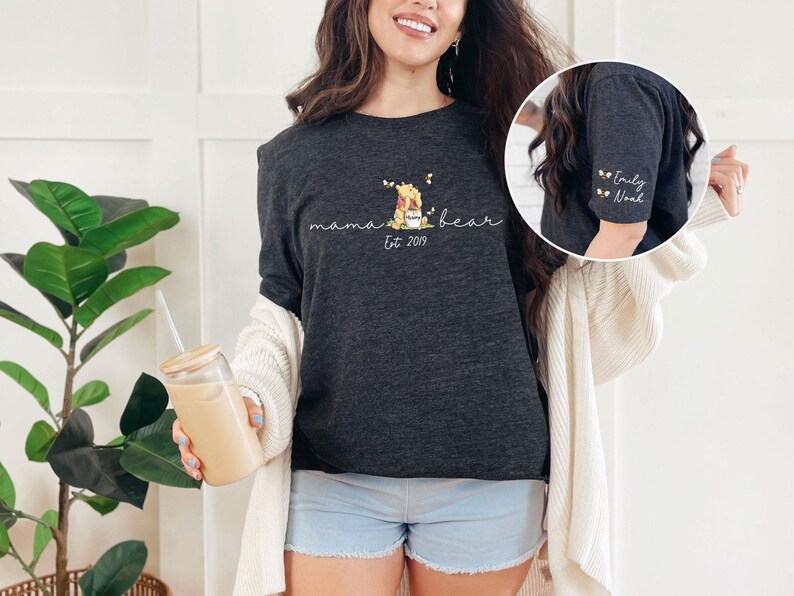 May include: A dark grey t-shirt with the text "mama bear est. 2019" and a small image of Winnie the Pooh holding a jar of honey. The sleeves have the names "Emily" and "Noah" embroidered on them.