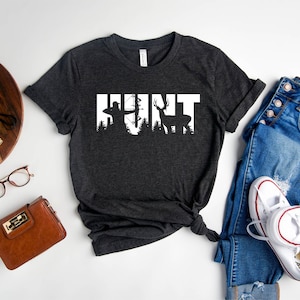 May include: Heather dark gray t-shirt with a white graphic of a hunter with a bow and arrow and a deer in the woods. The text "HUNT" is written in white letters.