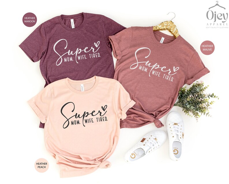 May include: Three t-shirts with the text "Super Mom Wife Tired" in different colors: heather maroon, heather peach, and heather mauve.