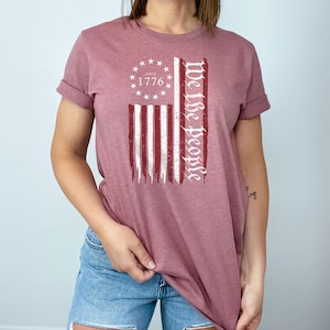 May include: A mauve t-shirt with a distressed American flag graphic and the text "We the People" printed in white. The flag graphic includes a circle with stars and the text "Since 1776".