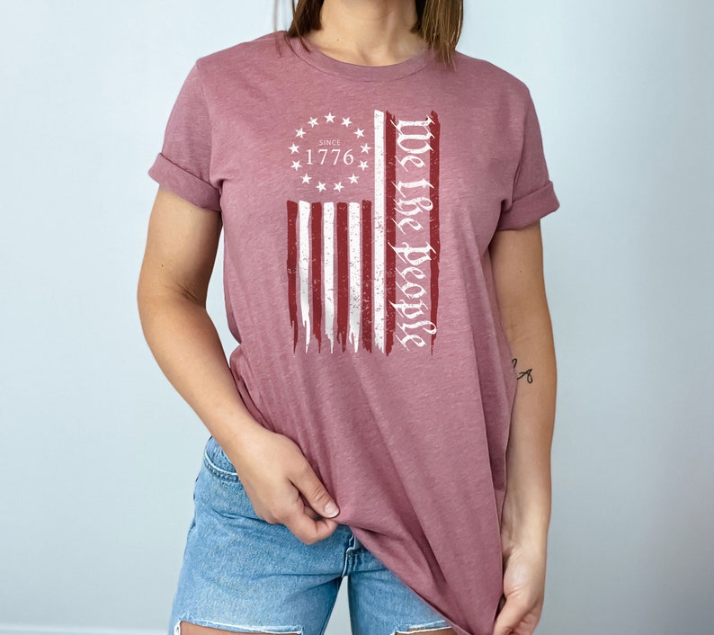 May include: A mauve t-shirt with a distressed American flag graphic and the text "We the People" printed in white. The flag graphic includes a circle with stars and the text "Since 1776".