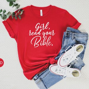 Pode incluir: Camiseta vermelha com o texto "Girl, read your Bible." em letras brancas. A camiseta est&aacute; dobrada e colocada sobre uma superf&iacute;cie branca com um par de cal&ccedil;as de ganga azuis e t&eacute;nis brancos.