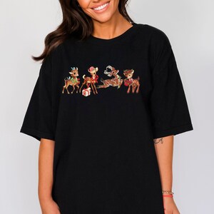 Pu&ograve; includere: Maglietta nera oversize con una grafica vintage di cinque renne vestite con abiti festivi. Le renne tengono dei regali e indossano dei fiocchi.