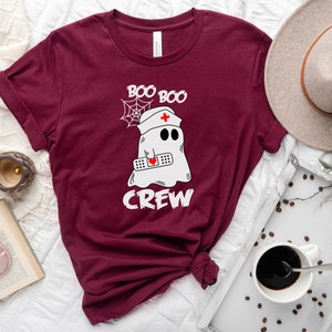 May include: Maroon t-shirt featuring a white graphic of a ghost wearing a nurse's hat and holding a bandage. The text "Boo Boo Crew" is printed on the shirt.