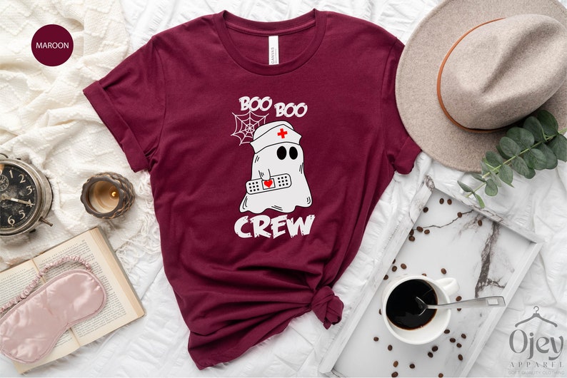 May include: Maroon t-shirt featuring a white graphic of a ghost wearing a nurse's hat and holding a bandage. The text "Boo Boo Crew" is printed on the shirt.