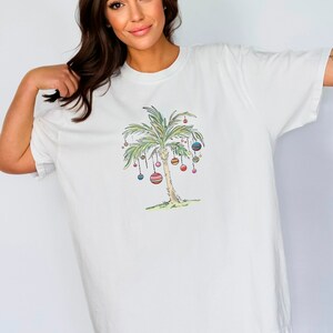 K&ouml;nnte beinhalten: Wei&szlig;es T-Shirt mit einem Aufdruck eines mit bunten Ornamenten geschm&uuml;ckten Palmenbaums. Der Palmenbaum ist im Cartoon-Stil gezeichnet.