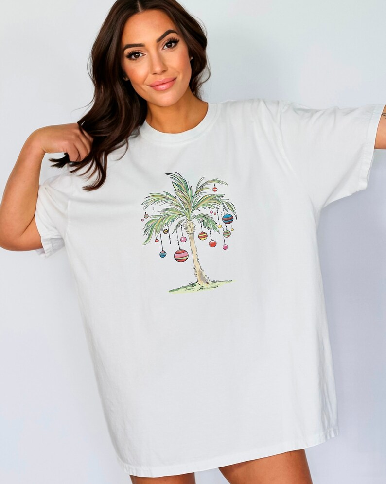 K&ouml;nnte beinhalten: Wei&szlig;es T-Shirt mit einem Aufdruck eines mit bunten Ornamenten geschm&uuml;ckten Palmenbaums. Der Palmenbaum ist im Cartoon-Stil gezeichnet.