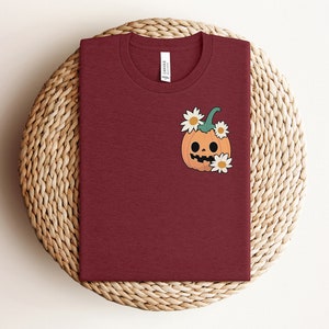 May include: A maroon t-shirt with a cartoon pumpkin with a smiling face and white flowers on the front.