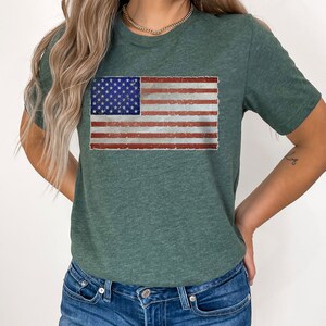 Pode incluir: Uma camiseta verde com um design de bandeira americana desbotada. A bandeira &eacute; vermelha, branca e azul.