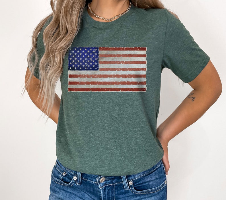 Pode incluir: Uma camiseta verde com um design de bandeira americana desbotada. A bandeira &eacute; vermelha, branca e azul.